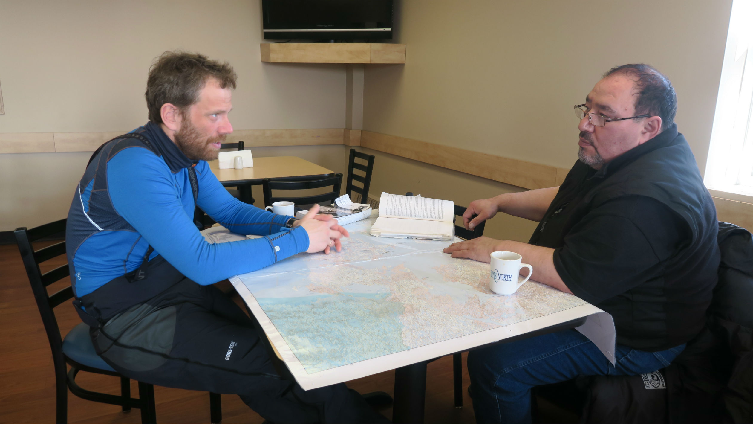 To menn rundt et bord med kart, bok og kaffekopp på.